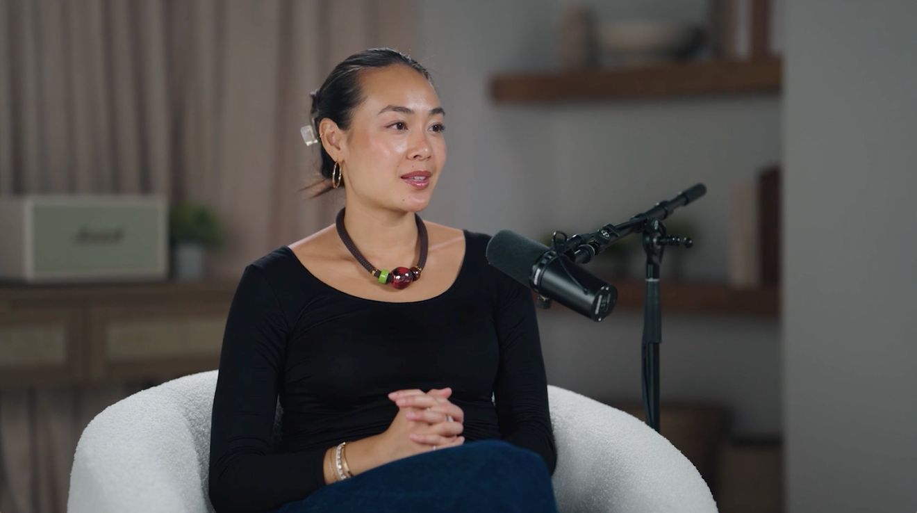 A woman in a black top and necklace seated in a white armchair at a podcast microphone