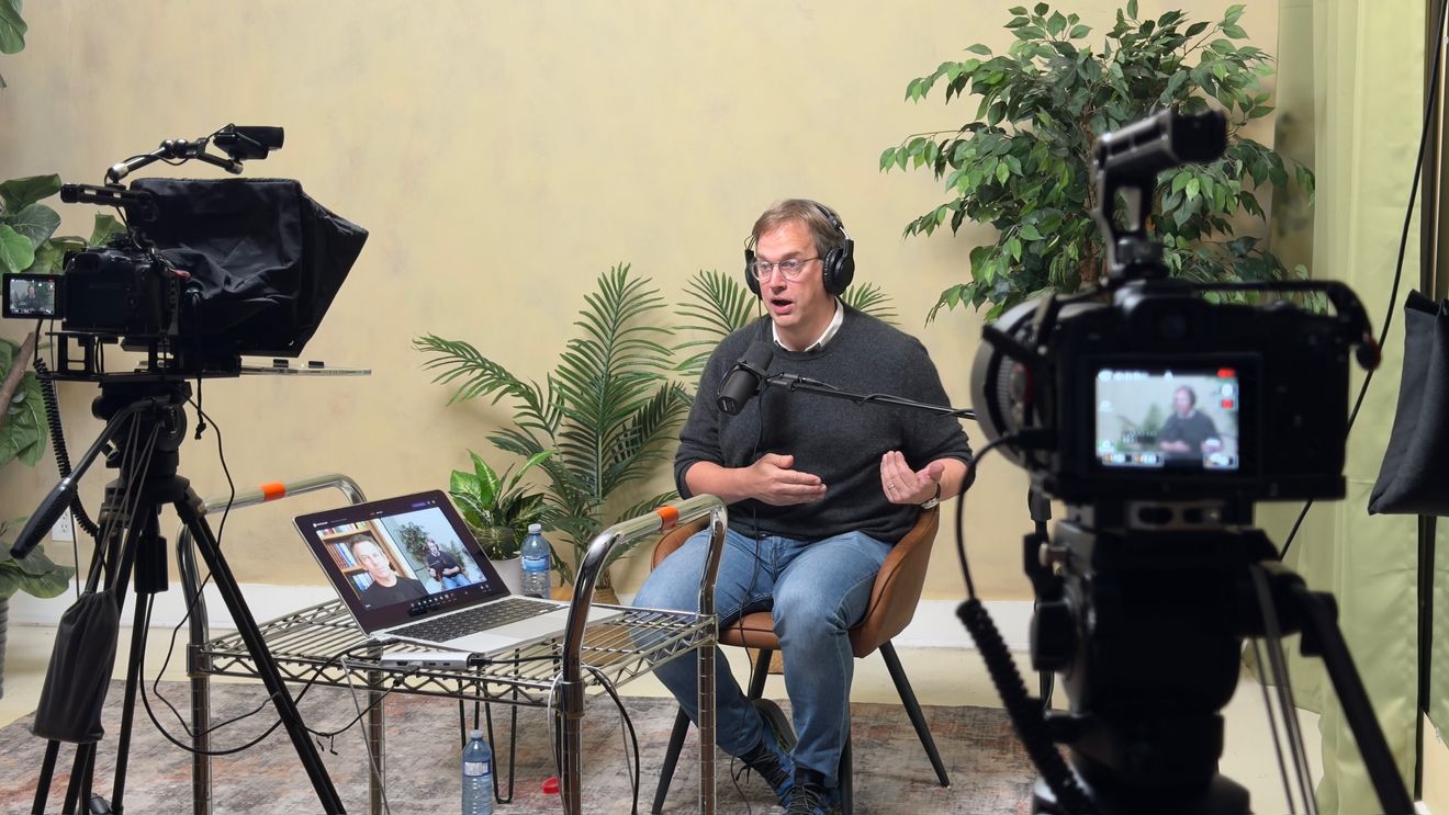 A man wearing headphones in a warm tan-toned room, looking toward a camera with a teleprompter